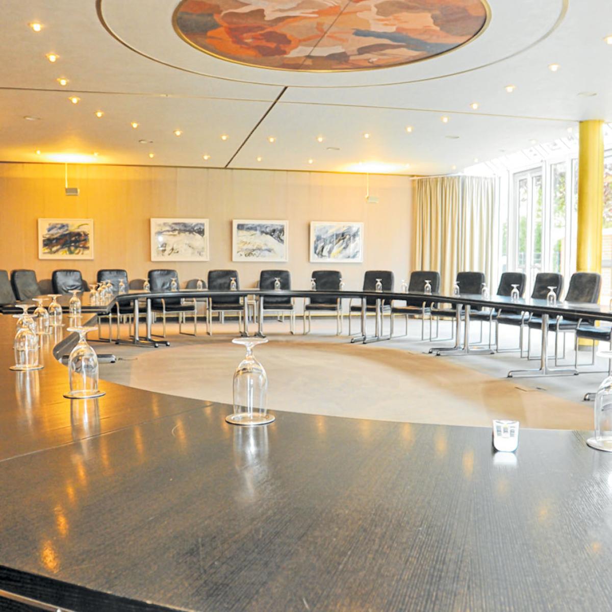 Raum mit rund aufgestellten Tischen auf denen an jedem Platz ein Trinkglas steht ein Sitzungssaal. 14 Lederstühle sind zu sehen.